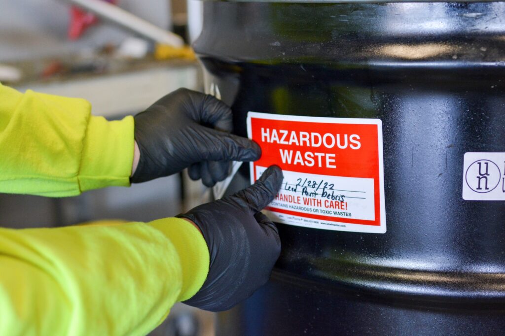 NEDT technician applies hazardous waste warning label to metal drum.